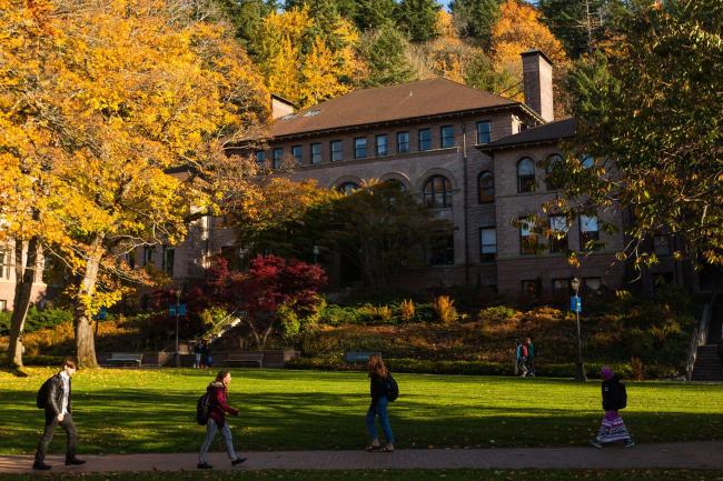 Students walk in front of a large brick building, with fall trees in the background.
